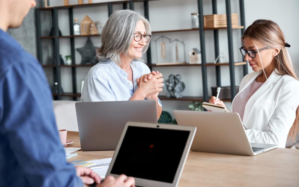 Smiling old female mentor training young interns at group office meeting.