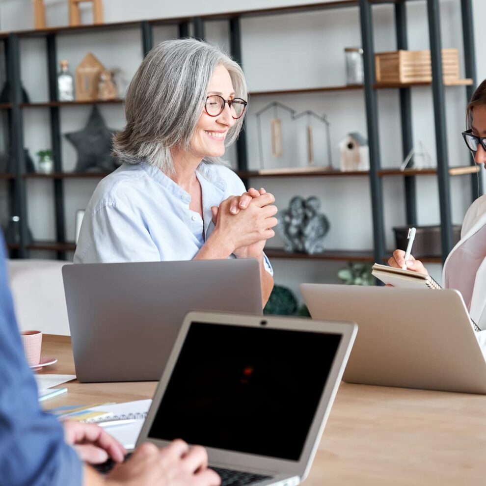 Smiling old female mentor training young interns at group office meeting.